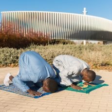 Two young senegalese men wearing traditional clothing are kneeling on prayer rugs, performing salat outdoors during the holy month of ramadan
